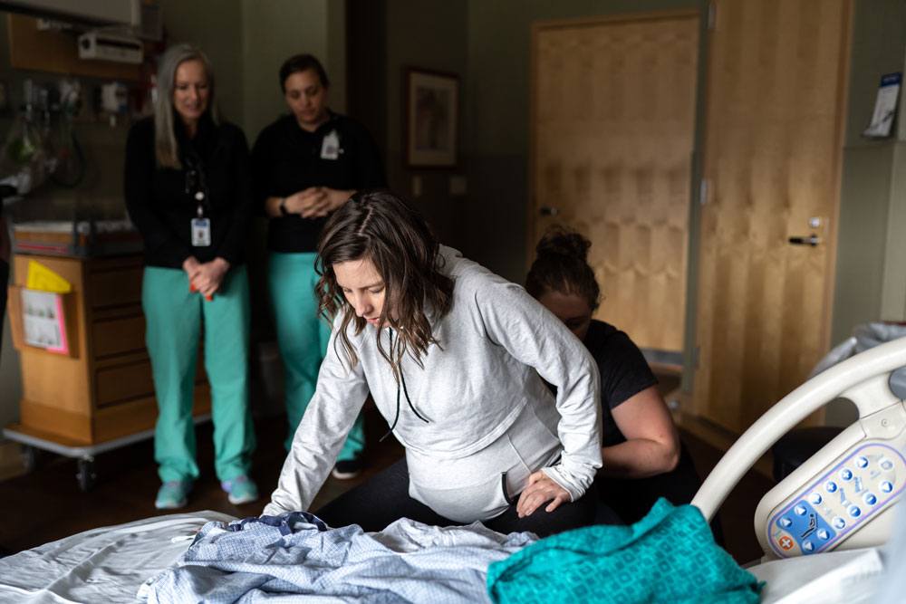 Woman leans on hospital bed while her doula does hip squeezes and her midwives watch from behind during a contraction at a birth in Utah