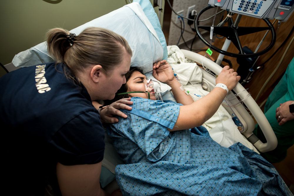 Mom lies in hospital bed with an oxygen mask on as a doula encourages her to take deep breaths during birth story