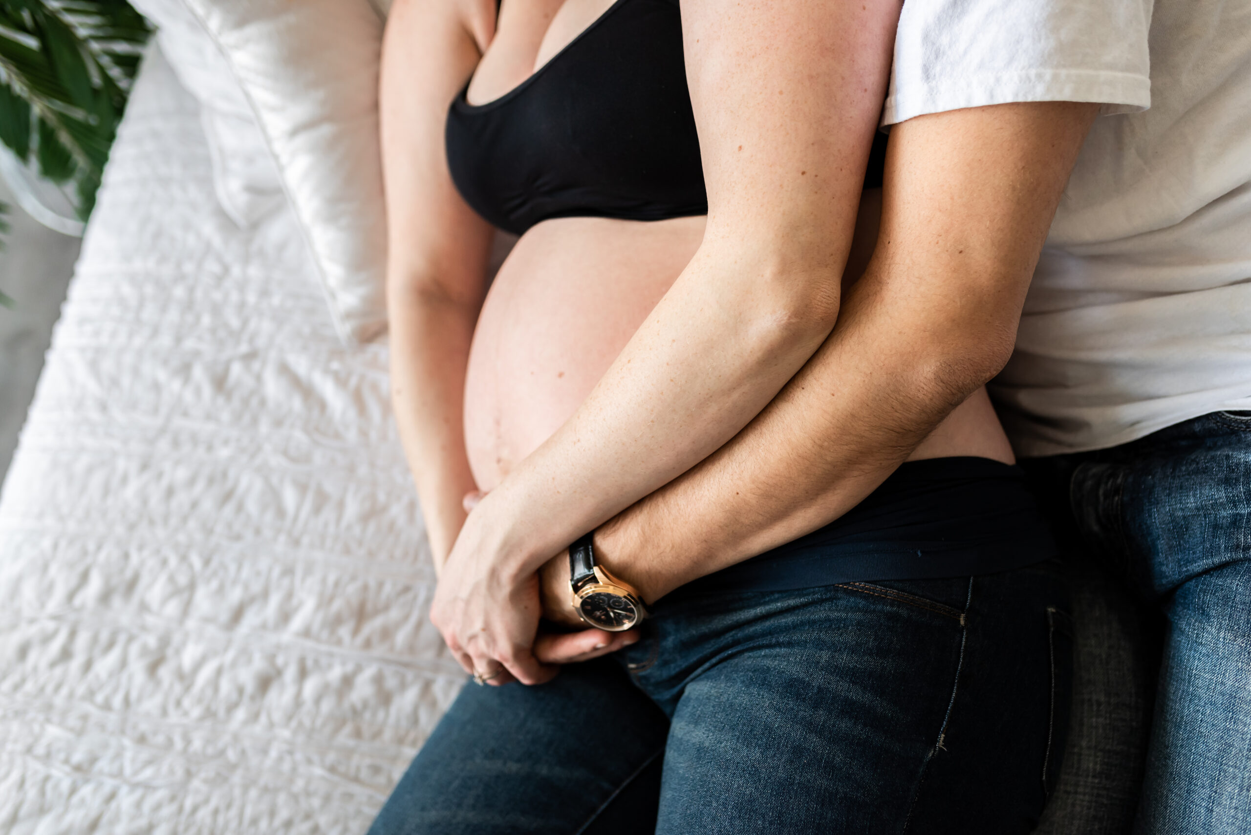 Husband and wife cuddle on a white bed while cradling her pregnant belly