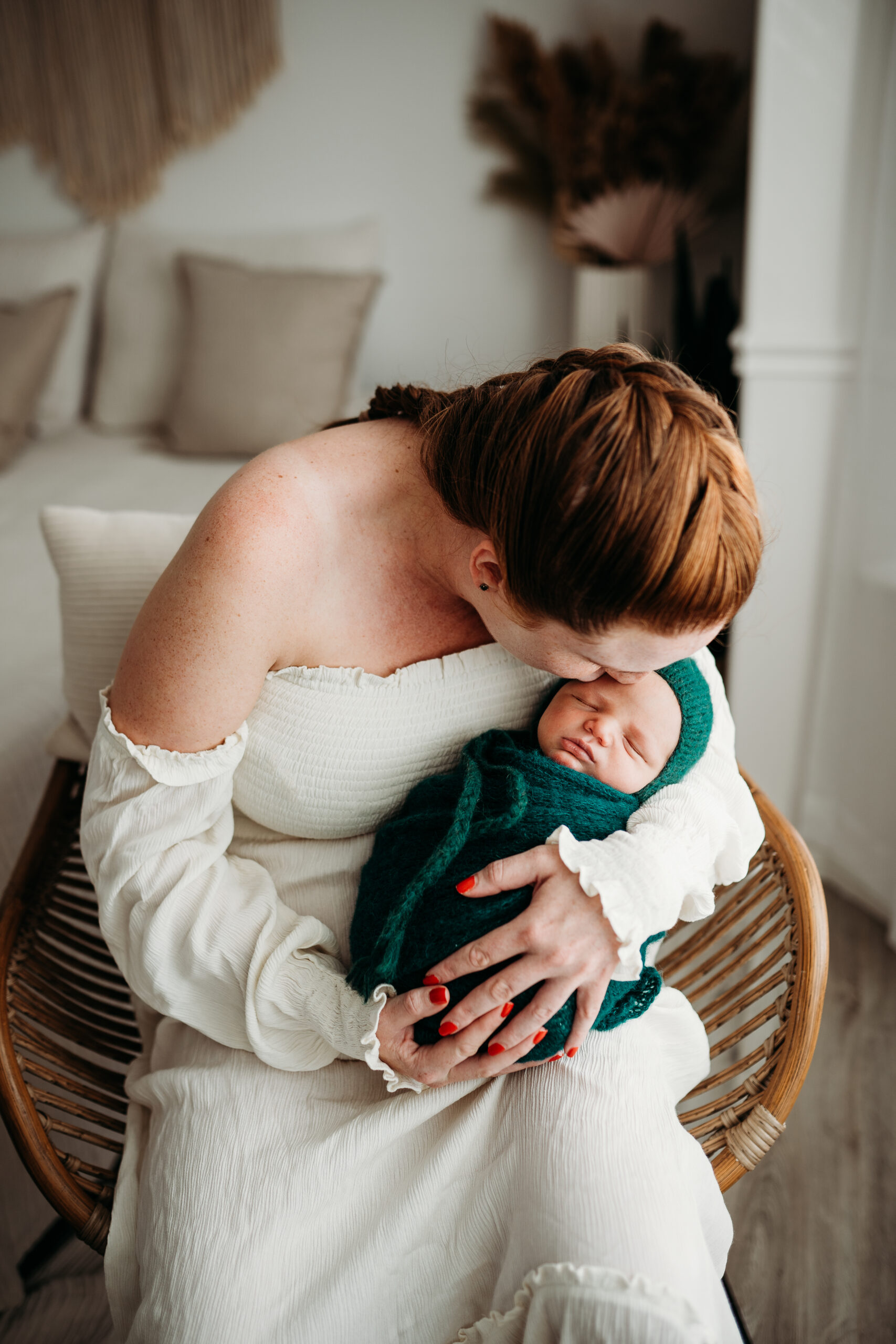 Mother holding her wrapped newborn baby while she kisses him sitting in a chair